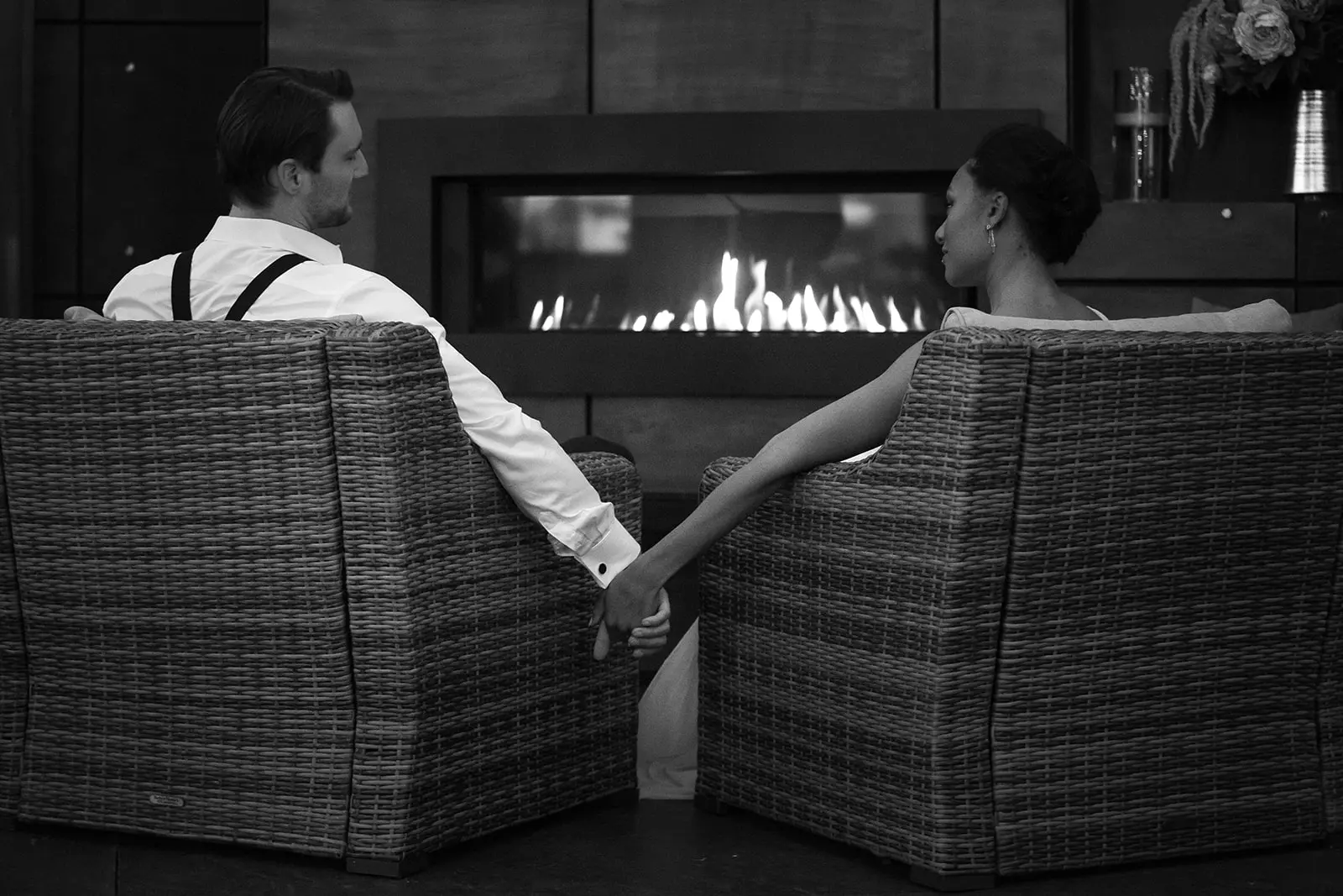 Black-and-white photo of a couple holding hands in wicker chairs facing the gas fireplace.