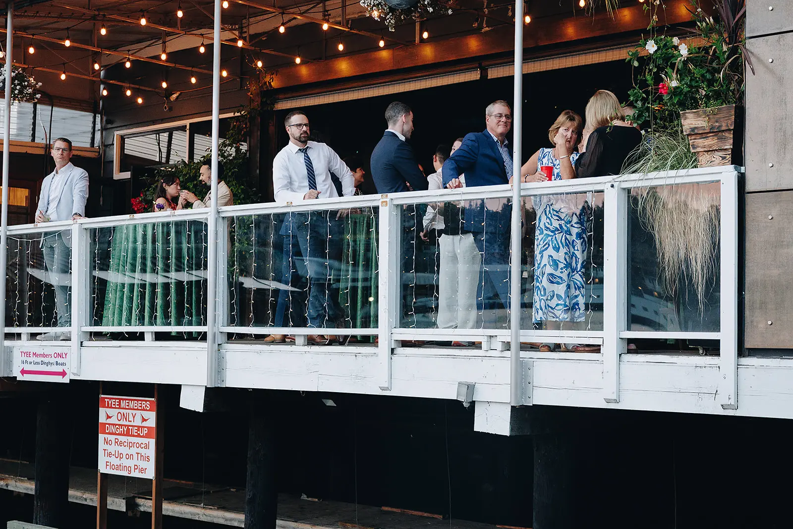 Guests talking and drinking on the covered patio railing above the Tyee Yacht Club dock, with string lights overhead.