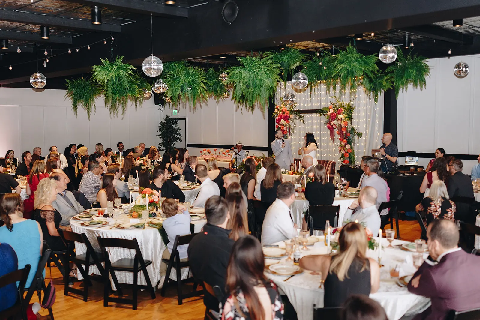 Guests seated at rounds in the main room during a toast, with hanging ferns and disco balls above and a floral arch at the front.